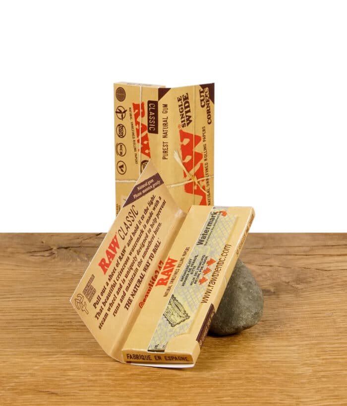 Zwei geöffnete Packungen RAW Classic Rolling Papers im Single Wide Format mit Cut Corners, auf einem Stein präsentiert